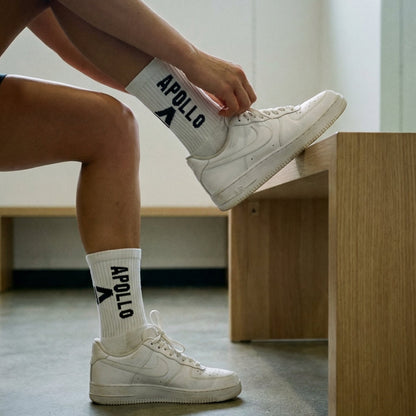 Person wearing white sneakers and socks with 'Apollo' branding, sitting on a wooden bench.